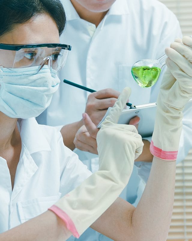 allegiance-industries-background-tall A woman examines a green liquid while wearing glasses, a mask and gloves