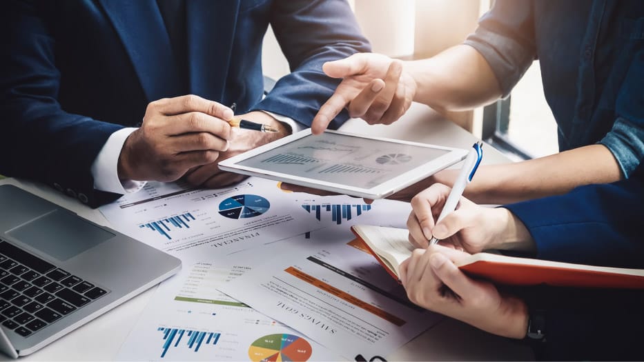 Businessmen and women point to graphs on paper and screens
